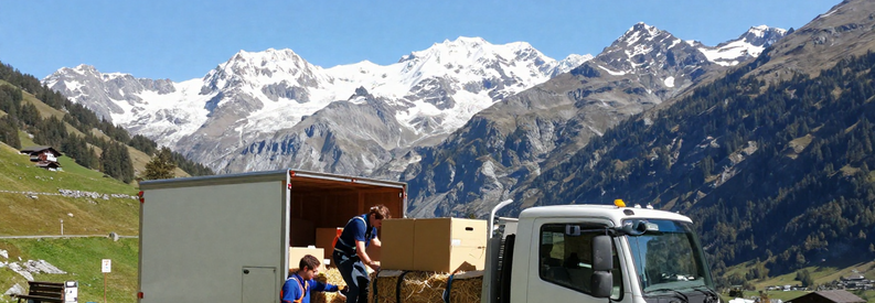 Camion déménagement négociant route sinueuse alpine Grisons, professionnels sécurisant chargement, vallée montagne