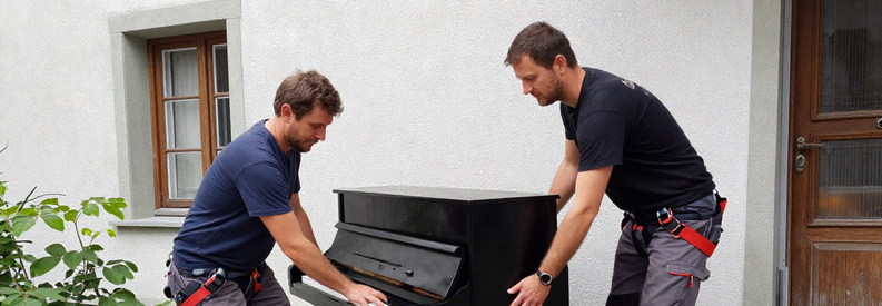 Deux déménageurs professionnels transportant un piano droit dans un escalier d'immeuble suisse avec sangles de levage